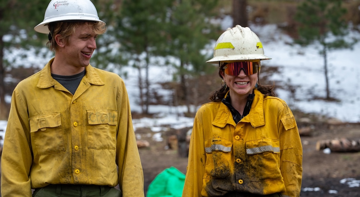 two students covered in soot and in wildfire fighting gear smile. Their helmets read "Colorado Firecamp."