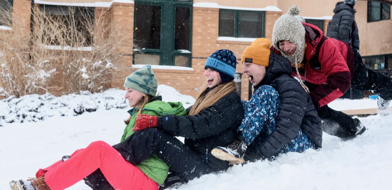 three students share a sled while a forth pushes them down a hill. All are laughing.
