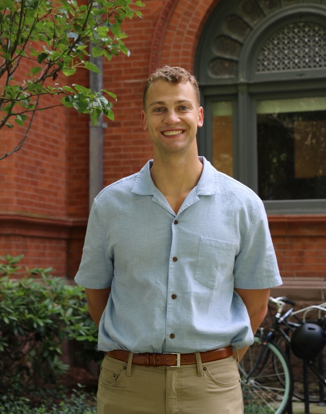 Owen Rask stands smiling with his hands clasped behind his back in front of a building on Yale's campus