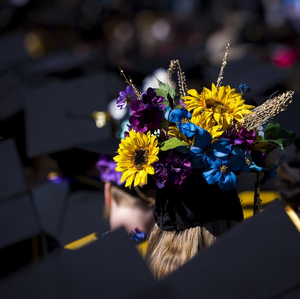 A person with blond hair and a graduation cap elaborately decorated with purple and gold flowers stands out in a sea of traditional black graduation regalia