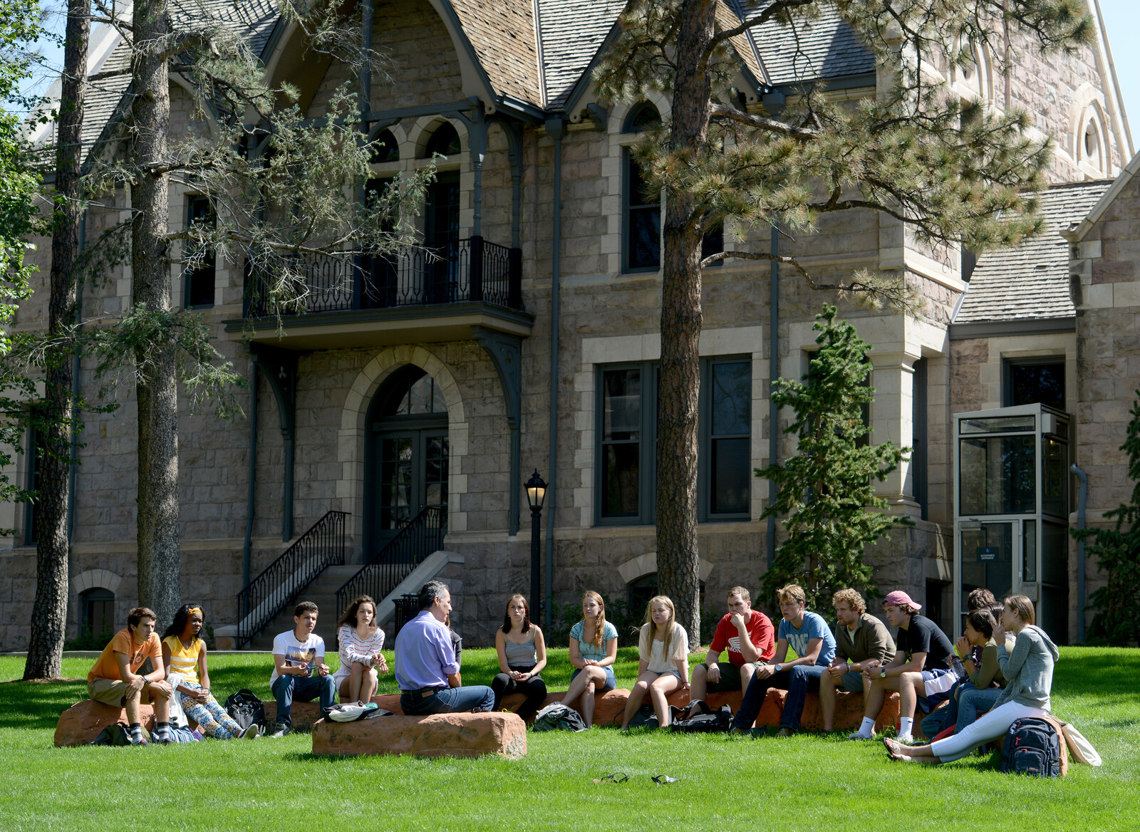 a class sitting on the lawn in Tava quad listening to a lecturer seated on a bench in front of Shove Chapel